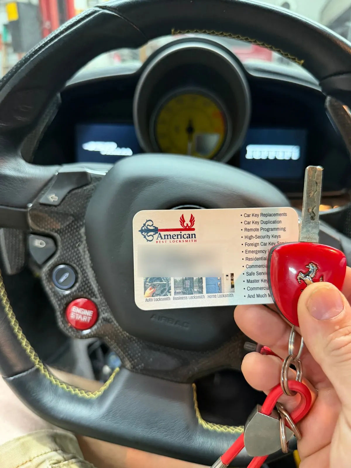 Ferrari key with red prancing-horse head and an American Best Locksmith business card held in front of the steering wheel and yellow tachometer of a 2013 Ferrari 458 Italia